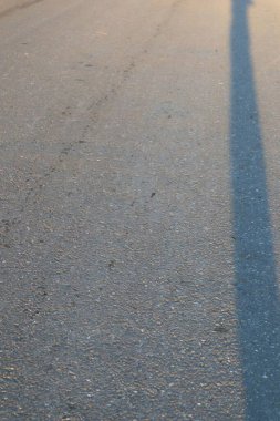 Güneş ışığı altında asfalt yol dokusuna yakın bir perspektif. Kumlu yüzeyi, ince gölgeleri ve monokromatik gri paleti vurgular, şehirsel alan duygusunu çağrıştırır..