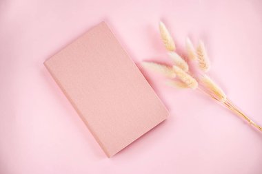 Yumuşak pembe bir defter, yumuşak bir pastel arka planda durur. Yanında da narin kurumuş çiçekler vardır. Not almak için mükemmel, davetkar ve sakin bir atmosfer yaratır..