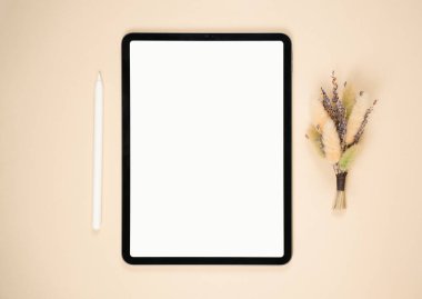Pürüzsüz bir tablet nötr bej bir yüzeyde, bir stilin yanında duruyor. Kurumuş çiçeklerden oluşan küçük bir düzenle tamamlanmış, sakin bir çalışma alanı ortamı yaratıyor..