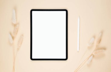 Boş bir tablet, bej bir arka planda stil ve zarif bir şekilde düzenlenmiş kurumuş bitkilerin yanında yer alır. Bu ayar, minimalist ve sanatsal bir çalışma alanını vurgular.