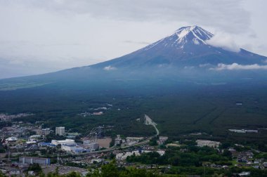Fuji Dağı, karla kaplı ikonik zirvesiyle, bulutlardan çıkar, doğanın görkeminin nefes kesici bir manzarasını sunar, yeşil manzaraların sakin bir zeminine karşı kurulur..