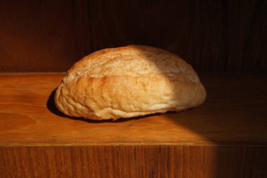 Yuvarlak bir somun taze pişmiş ekmek, sıcak güneş ışığıyla aydınlatılmış ahşap bir rafta oturur ve rahat bir kafede davetkar ve iştah açıcı bir atmosfer yaratır.