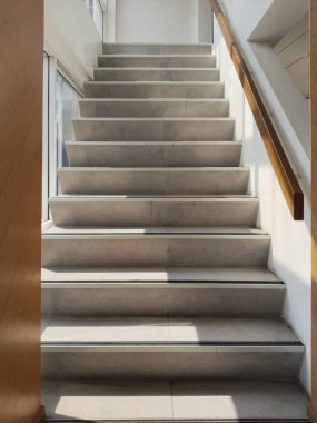 Büyük pencerelerden içeri güneş ışığı süzülen, merdivenlere gölge düşüren modern bir kapalı merdiven. Merdivenlerin bir tarafında ahşap korkuluklar olan parlak bir tasarımı var..