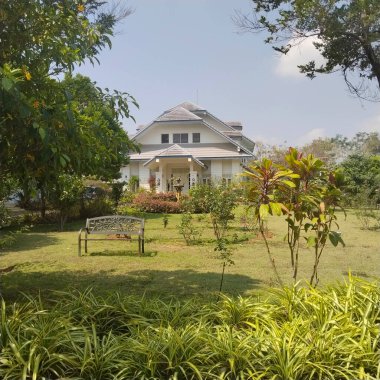 Yeşillik ve bakımlı bir bahçeyle çevrili büyüleyici bir ev. Ev eğimli bir çatı ve ön planda bir bank bulunan bir karşılama sundurmasına sahip..