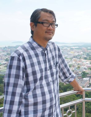 Kareli tişörtlü bir Asyalı gözlüklü arka planda tepeler olan bir balkonda duruyor..