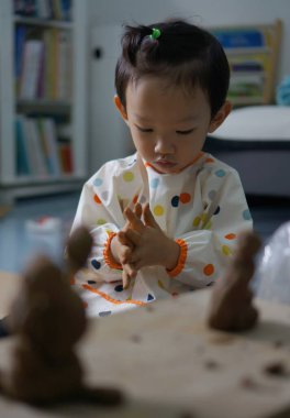 A young child focused on playing with clay, engaged in the process of creation. The child is seated at a wooden table, molding the clay with focus and care