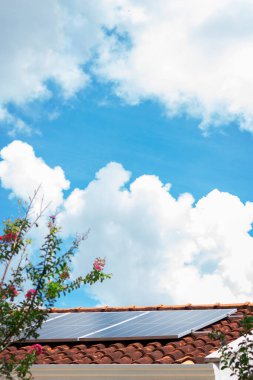 Güneşli ve bulutlu bir günde çatıdaki güneş panelleri. Fotovoltaik Kurulum Etrafında Yapraklar ve Çiçekler Olan Bir Evde.