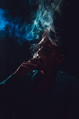 Siyah Gözlüklü Genç Adam Siyah Arkaplanda Dumanlı Puro İçiyor.