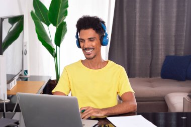 Black Man Wearing Yellow T-shirt at Remote Job in His House. He is Happy in a Teleworking Meeting at Desk With Laptop clipart