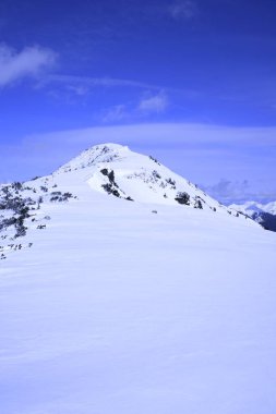 Berrak mavi bir gökyüzünün altında karla kaplı bir dağ zirvesinin yer aldığı sakin bir kış manzarası. Ön taraf taze karla kaplanmış. Huzurlu ve sakin bir manzara yaratıyor..