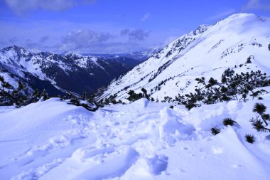 Açık mavi bir gökyüzü altında karla kaplı dağları barındıran nefes kesici bir kış manzarası. Ön planda el değmemiş ayak izleri var. Uzak tepeler kısmen bulutlarla örtülü..