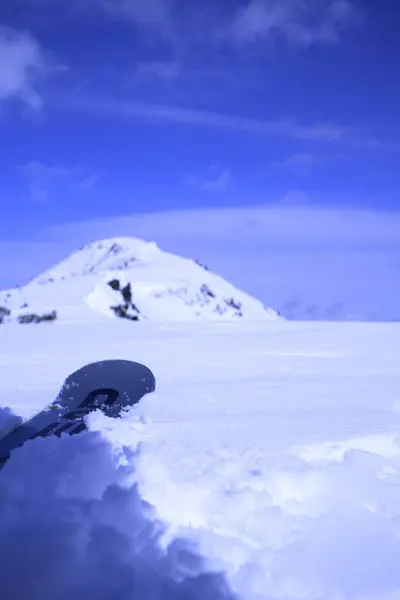 Arkasında dağ olan karlı bir manzara. Ön planda kısmen kara gömülü bir kayak ya da snowboard yer alıyor, açık mavi gökyüzünün altında bazı bulutlar var..
