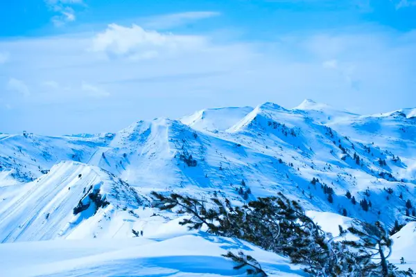 Berrak mavi gökyüzünün altında karla kaplı dağları barındıran sakin bir kış manzarası. Sahne, doğanın güzelliğini, yumuşak yamaçlar ve uzak tepelerle yakalıyor ve sakin bir atmosfer yaratıyor..