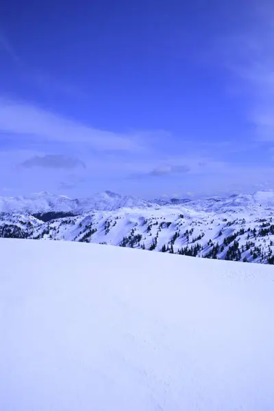 Berrak mavi bir gökyüzünün altında engin, karlı bir manzara, kayan tepeler ve karla kaplı uzak dağlar. Sahne bir sükunet ve kış güzelliği hissi veriyor..