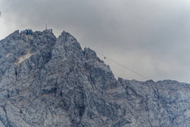 Bulutlu bir gökyüzünün altında engebeli bir dağ zirvesi, zirvede bir teleferik ve bir bina var. Kayalık arazide keskin kayalıklar ve dramatik bir manzara sergileniyor..