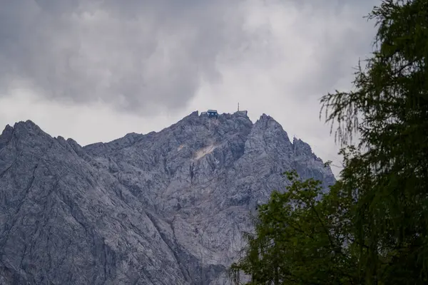 Bulutlu bir gökyüzünün altında engebeli tepeleri olan dramatik bir dağ manzarası. Zirvede kayalık arazi ve ön planda yeşillik ile çevrili küçük bir bina görülebilir..