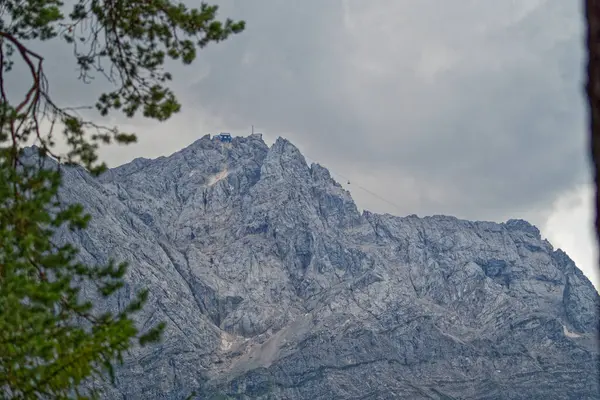 Bulutlu bir gökyüzünün altındaki engebeli bir dağ zirvesinin dramatik bir görüntüsü, bir teleferik hattı görünür. Kayalık arazi, karanlık bulutların kontrastı ile vurgulanır ve karamsar bir atmosfer yaratır..