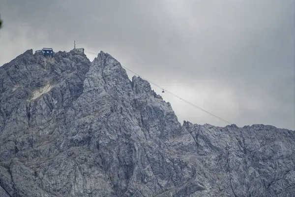 Bulutlu bir gökyüzünün altındaki engebeli bir dağ zirvesi. Zirvede bir binaya giden teleferik hattı var..