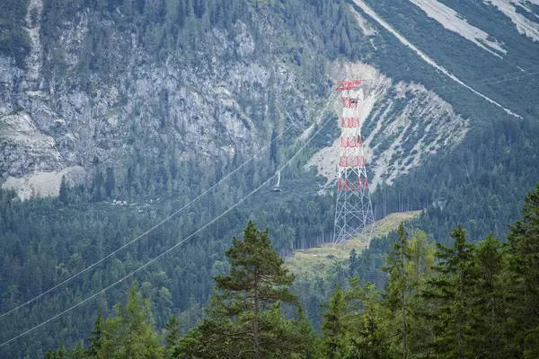 Bir teleferik sistemi ve şanzıman kulesinin yer aldığı dağlık bir manzaranın manzarası. Ön planda yemyeşil ağaçlar yer alırken, arka planda dik, kayalık yamaçlar ve sık ormanlar yer alıyor..