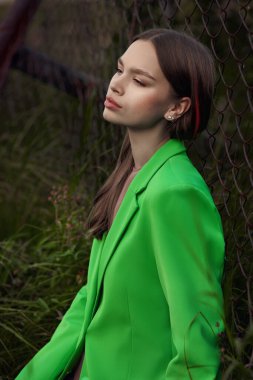 Göl kenarındaki çimlerde dinlenen genç bir kadın. Bej elbiseli, yeşil ceketli, şehvetli bir kadın.