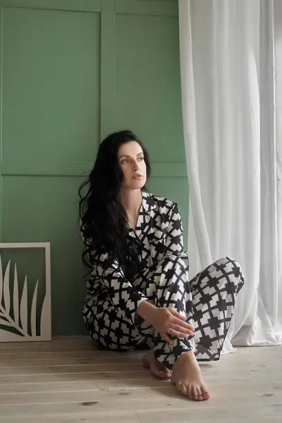 Woman in pajamas is sitting on the floor of the house. Long black brunette hair