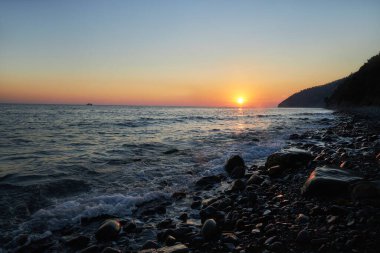Güneş ufkun altına batıyor, kayalık kıyı şeridi, doğa güzelliği huzur içinde. Dalgalar yavaşça kıyıya vururken, siluetli kayalıklar, koyu gökyüzüne karşı görkemli bir şekilde duruyor.