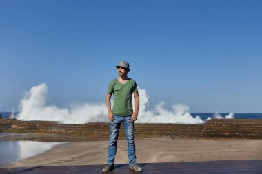 Sıradan giyinmiş bir adam, mavi bir gökyüzünün altında arka planda kırılan büyük dalgalarla bir rıhtımda duruyor. Tenerife, İspanya