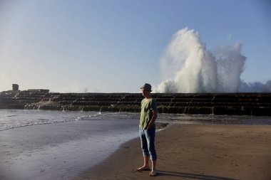 Şapkalı yalnız bir adam sakin bir günbatımında dalgaların heybetli kıyılarla çarpışmasını gözlemler. Tenerife, İspanya