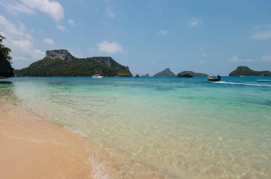 Turkuaz sular, güneşli bir günde arka planda yeşil tepeler ve teknelerle dolu kumlu bir plajla karşılaşır.