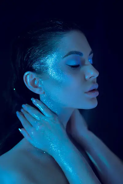 stock image Woman with shimmering blue cosmetic makeup, serene pose, face and hand glowing under a soft blue light