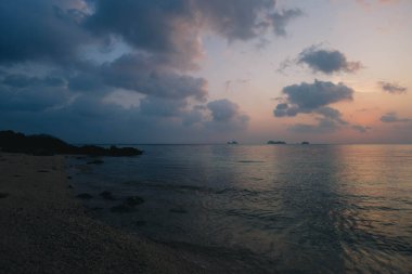 Sakin bir sahilde bulutların arkasında güneş batarken huzurlu bir akşam. Suya yumuşak bir parıltı yayıyor.