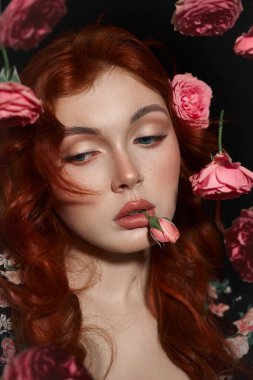 Kırmızı saçlı, mavi gözlü bir kadın başka tarafa bakıyor, pembe güllerle çevrili.