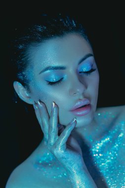 Beauty Woman covered in shimmering substance under blue lighting touches her face gently, makeup cosmetic