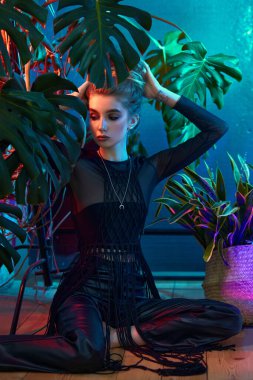 Genç bir kadın canlı tropikal bitkilerle çevrili, siyah deri kıyafetler giyiyor ve uzun kollu, katıksız bir bluz giyiyor. Neon ışığı parlıyor