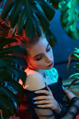 Genç bir kadın canlı tropikal bitkilerle çevrili, siyah deri kıyafetler giyiyor ve uzun kollu, katıksız bir bluz giyiyor. Neon ışığı parlıyor