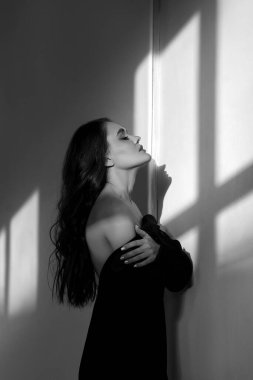 Uzun siyah saçlı kadın beyaz duvara yaslanıyor, güneş ışığı pencereden içeri akıyor ve gölgeler bırakıyor. Siyah bir ceket giyiyor ve yüzü yukarı doğru eğik.