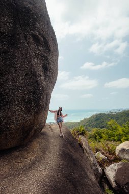Bir kadın, arkasında devasa bir kaya formasyonuyla bir uçurumun kenarında yürüyor. Tropikal ada okyanus manzarası ve uzaktaki yemyeşil bitki örtüsü sunar..