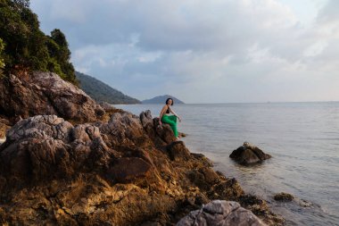 Gün batımında kayalık bir sahilde oturan bir kadın sakin denizi seyrediyor. Bereketli yeşillik ve uzak bir dağ sırası pitoresk bir zemin sağlar..