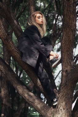 Siyahlar giymiş genç bir kadın gündüzleri bir ağaçta oturuyor. O, yüzünü çerçeveleyen uzun sarı saçlarıyla kameradan uzağa bakıyor..