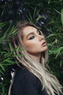 Sarışın saçlı güzel kadın yeşil yaprakların arasında poz veriyor, yüzü kısmen yapraklarla gizlenmiş. Siyah bir kazak giyiyordu, gözlerini dikip, gizemli bir hava yaratıyordu.