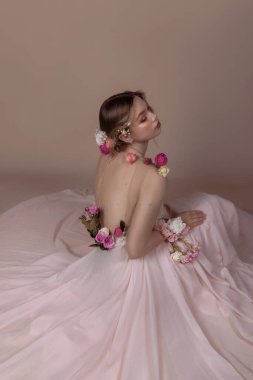Çiçeklerle süslenmiş pembe bir elbise giyen bir kadın stüdyo ortamında yerde oturuyor..