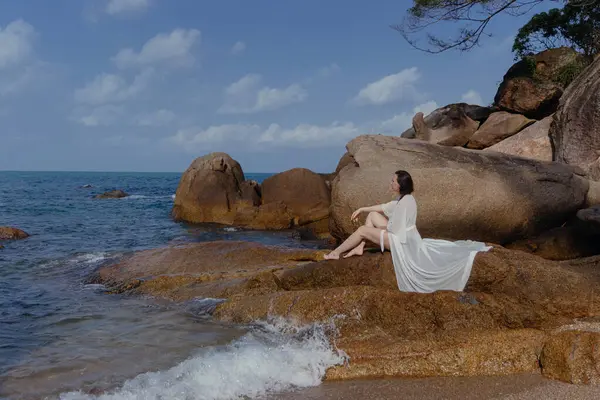 Stock image Woman in white robe sits on rock by calm blue ocean, legs outstretched towards water. White clouds in sky, seems content in peaceful view.