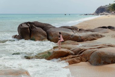 Tayland 'da bir plajda mayo giymiş bir kadın kayalık bir çıkıntıda duruyor ve okyanus dalgaları onu çevreleyip geçiyor..