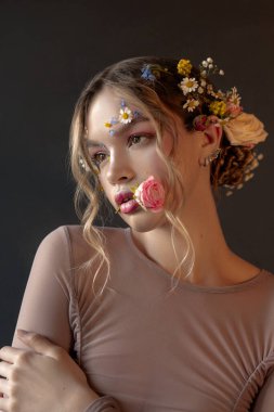 Uzun sarı saçlı, çiçekle süslenmiş genç bir kadın. Yan tarafa bakıyor. Yüzü de narin çiçeklerle süslenmiş..