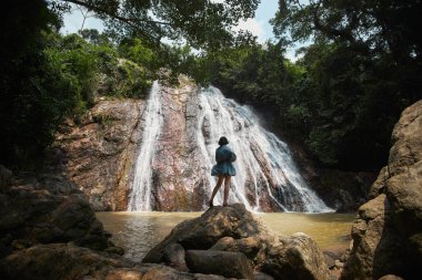 Yoğun bir ormanda çağlayan bir şelaleye bakan bir kayanın üzerinde duran kadın. Şelale, yemyeşil yemyeşil bir kayalıklardan aşağı akar..