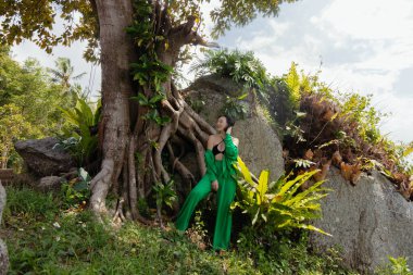 Kadın tropik bir ortamda büyük bir ağacın yanında poz veriyor. Yeşil bir elbise giyiyor ve etrafı yemyeşil yapraklar ve kayalarla çevrili..