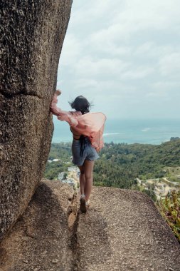 Bir kadın dar bir uçurum kenarında yürüyor, sırtı kameraya dönük, arkasında mavi okyanus ve tropik yeşillik ile..