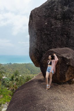 Bir kadın tropik bir adanın kayalık tarafında, büyük ve dengesiz bir kayanın altında duruyor. Okyanusa ve aşağıdaki yemyeşil ormana bakıyor..