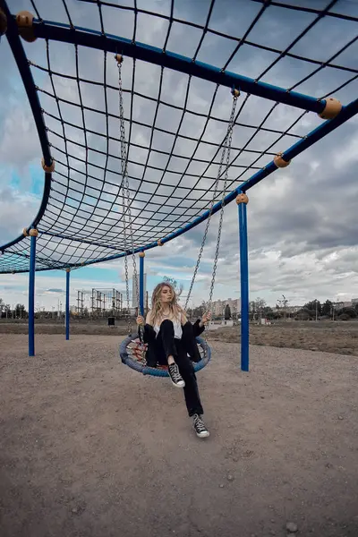 stock image A young woman sits on a swing at a playground, looking off into the distance. The sky is cloudy, and the playground is empty.