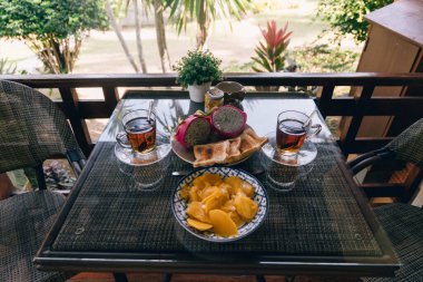 Balkonda taze mango, hamur işi ve çay servis edilen enfes bir tropikal kahvaltı. Manzara parlak ve davetkar, canlı bitkiler ve yumuşak sabah ışığı ile çevrili..